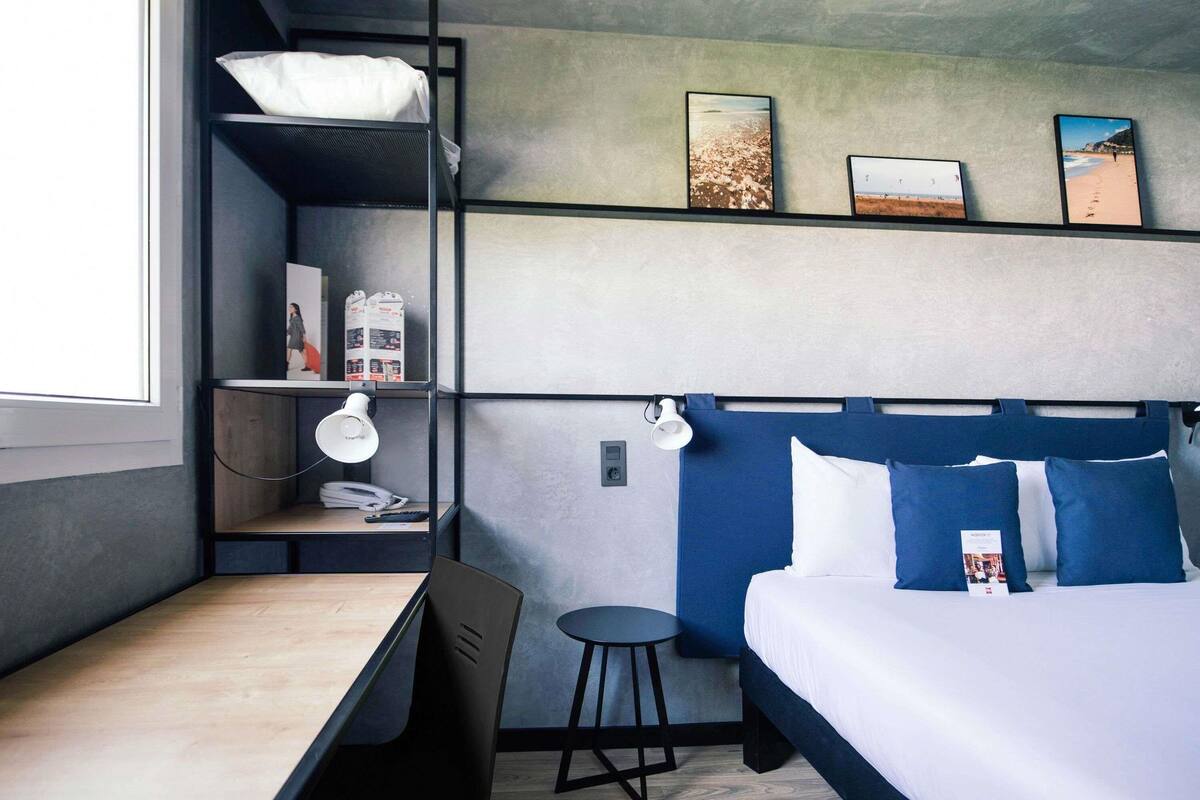 A modern hotel room with a bunk bed, a desk with a chair, a small stool, and a bedside table.