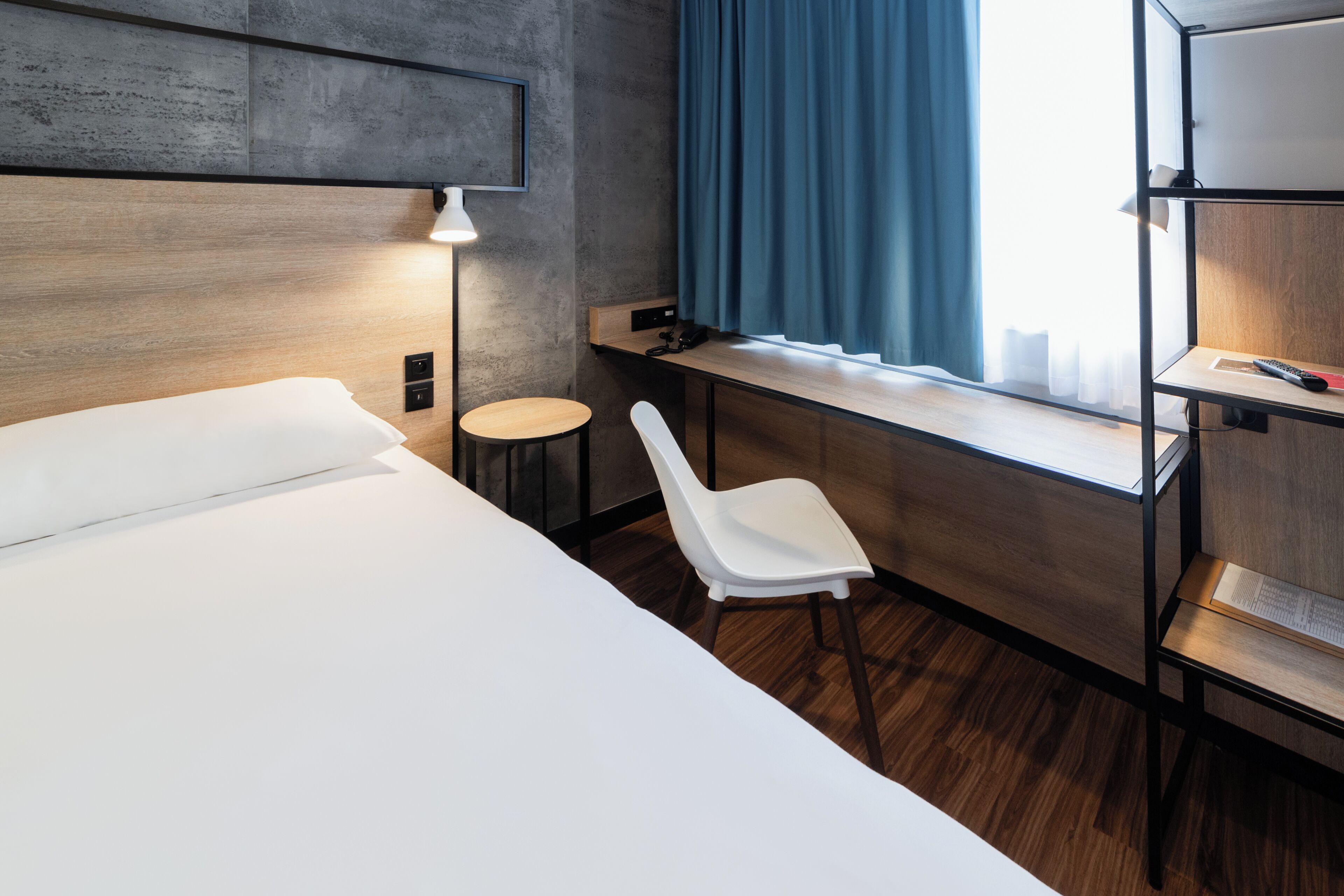 A hotel room with a bed, a desk, a chair, a lamp, and a window with curtains.