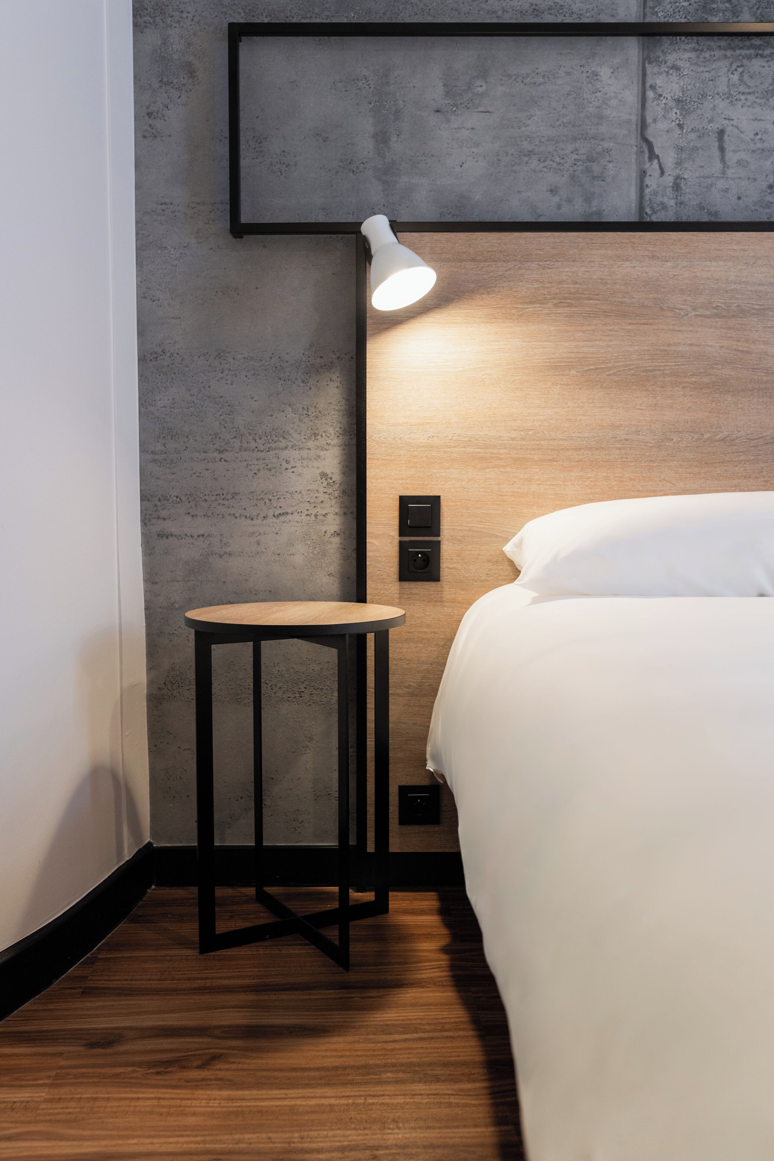 Una habitación de hotel moderna con suelo de madera, una mesita redonda pequeña, una cama con ropa blanca y una lámpara de pared.