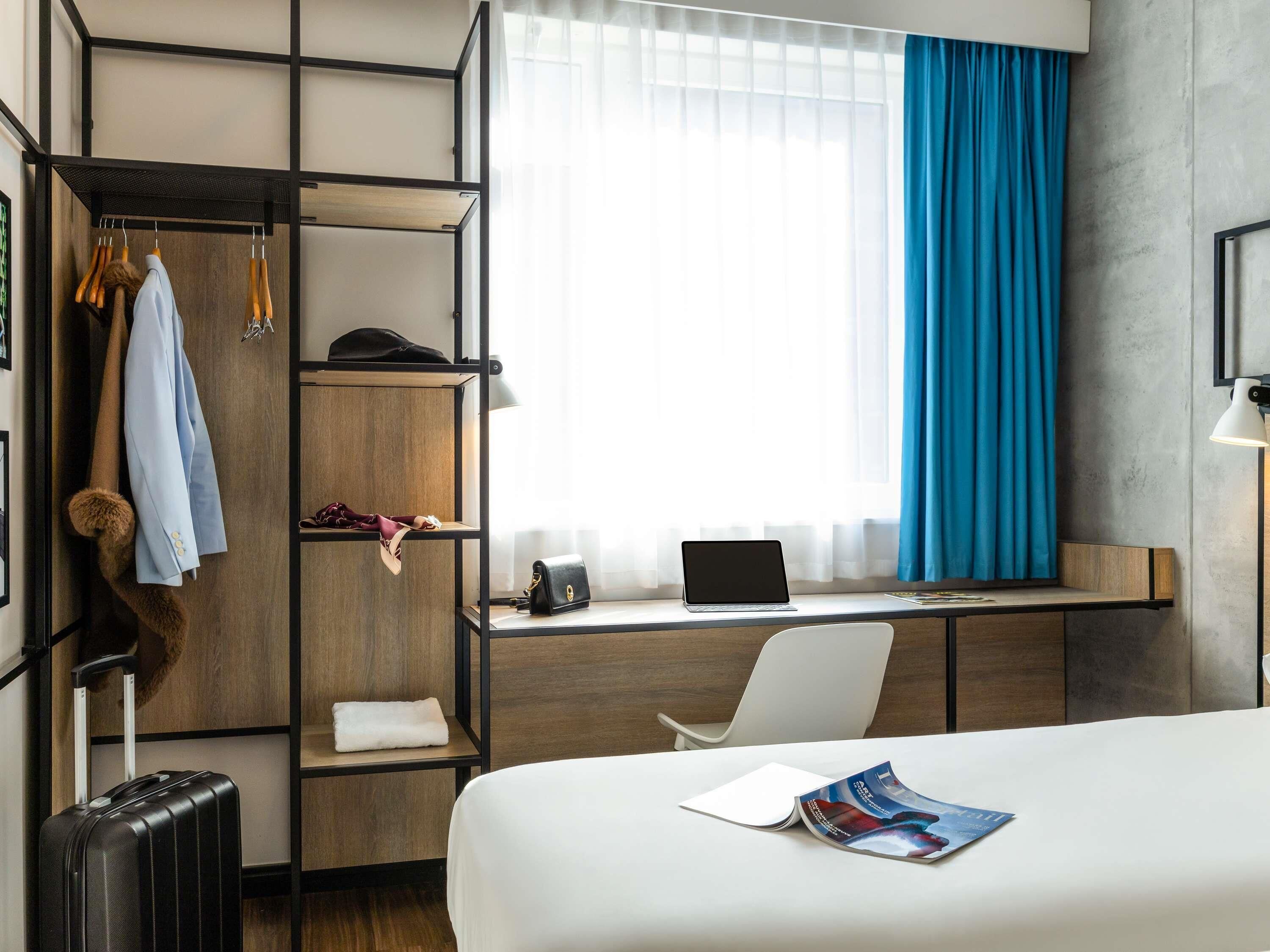 Habitación de hotel con cama, escritorio con computadora port átil, silla, armario con ropa y maleta.
