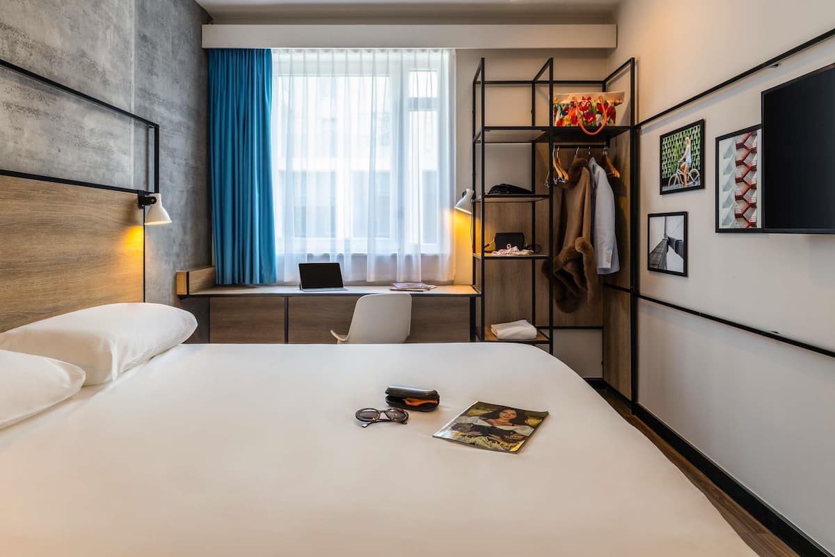 A hotel room with a bed, a desk with a laptop, a chair, a wardrobe, and a window with curtains.