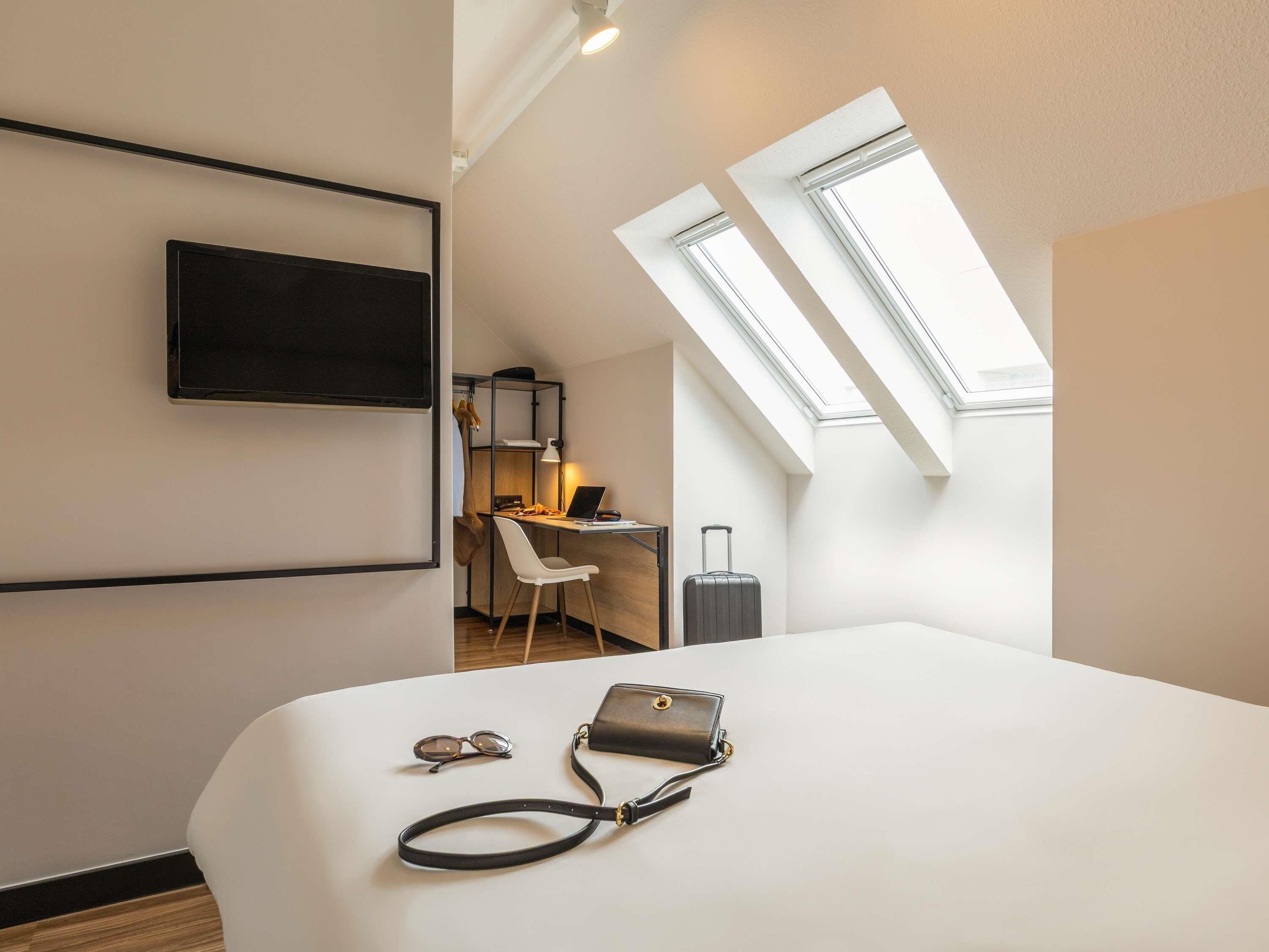 Une chambre d’hôtel comprenant un lit, un bureau avec une chaise, une télévision et deux fenêtres de toit.