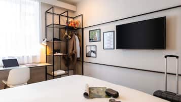 Una habitación de hotel con una cama, un escritorio con una computadora portátil, un televisor y un armario con ropa y un bolso.