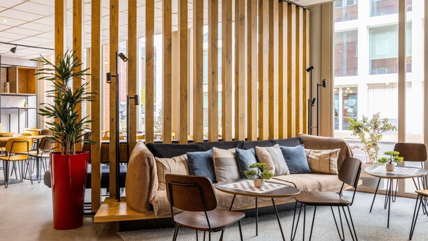 Un área de descanso moderna con un separador de madera curvada, un sofá con varios cojines y una mesa redonda con sillas.