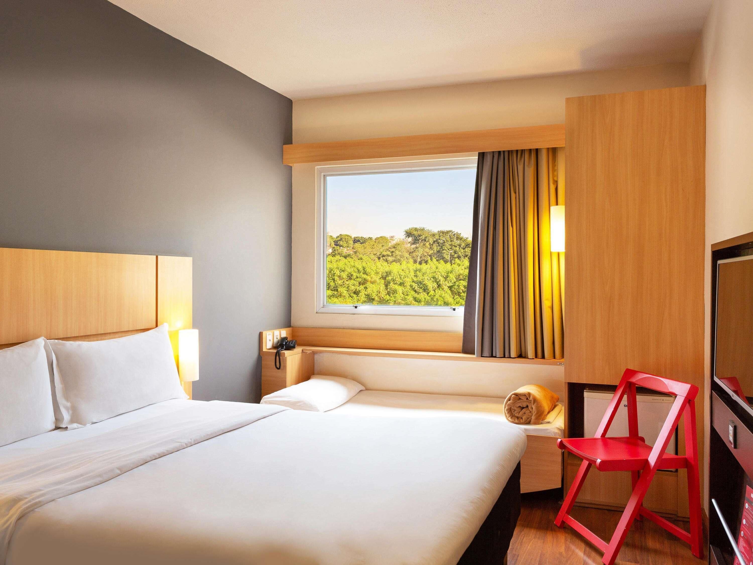 Habitación de hotel con cama, escritorio con silla, ventana y vista a vegetación, y televisor.