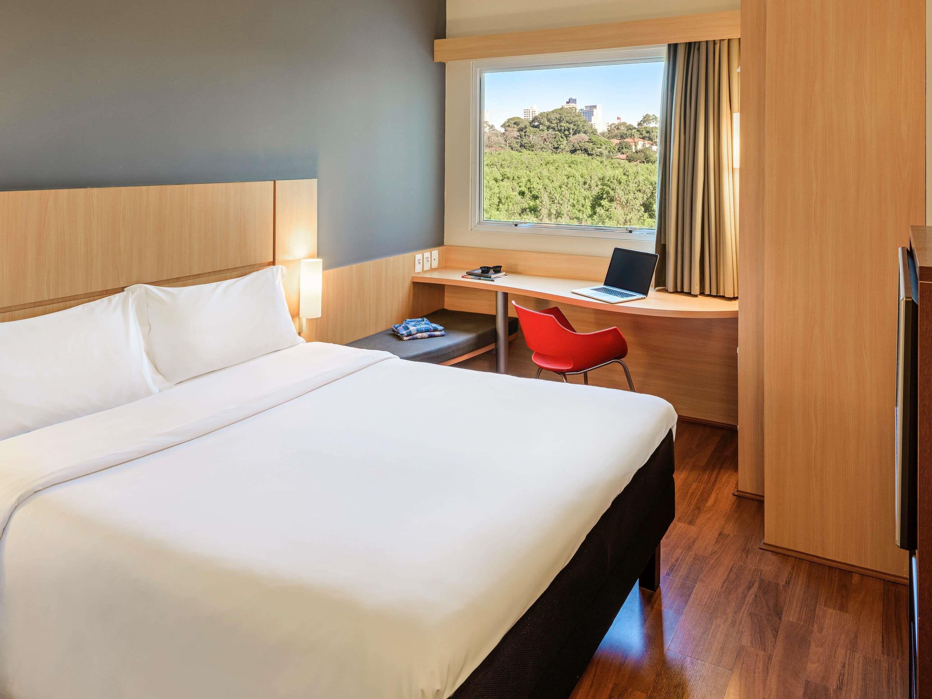 A hotel room with a large bed, a desk with a laptop, a red chair, and a view of a cityscape through a window.