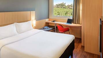 A hotel room with a large bed, a desk with a laptop, a red chair, and a view of a cityscape through a window.