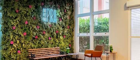 Una habitación con una pared verde cubierta de follaje y flores, un banco, una silla, una mesita y un ventanal grande.