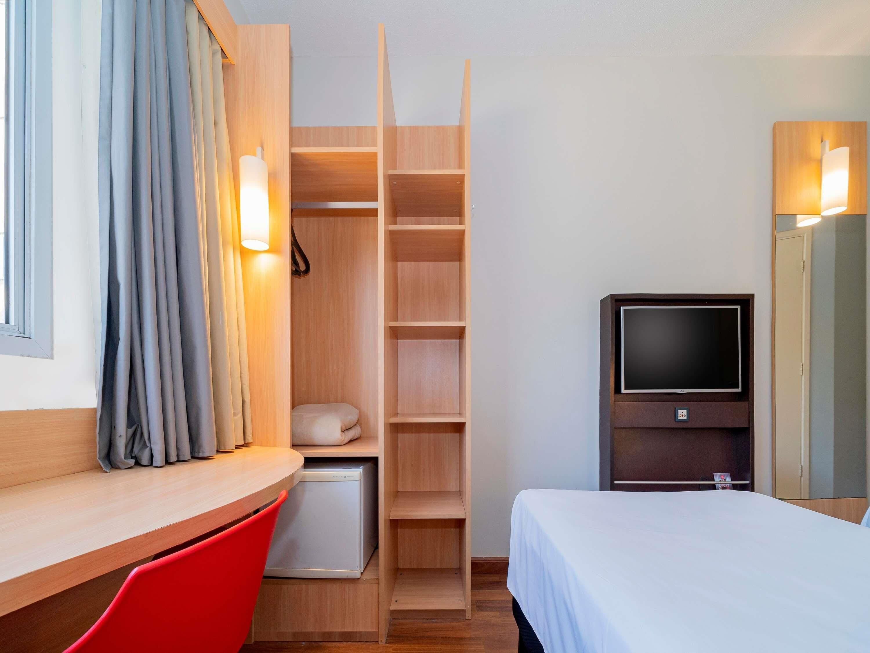 Habitación de hotel con cama, televisión, escritorio con silla, armario y un refrigerador pequeño.