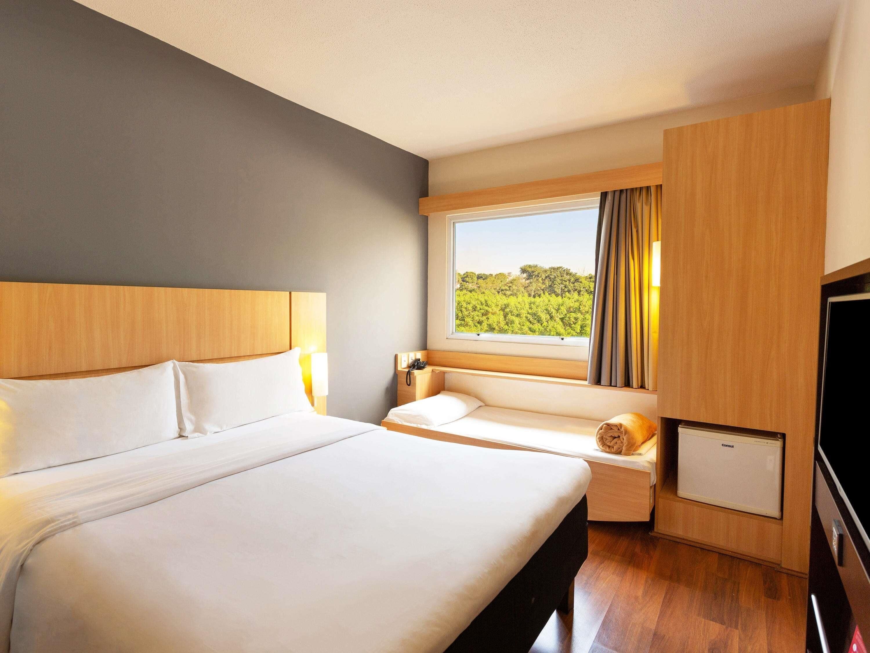 A hotel room with a large bed, a bedside table with a lamp, a small desk with a chair, a window with a view of greenery, and a minibar.