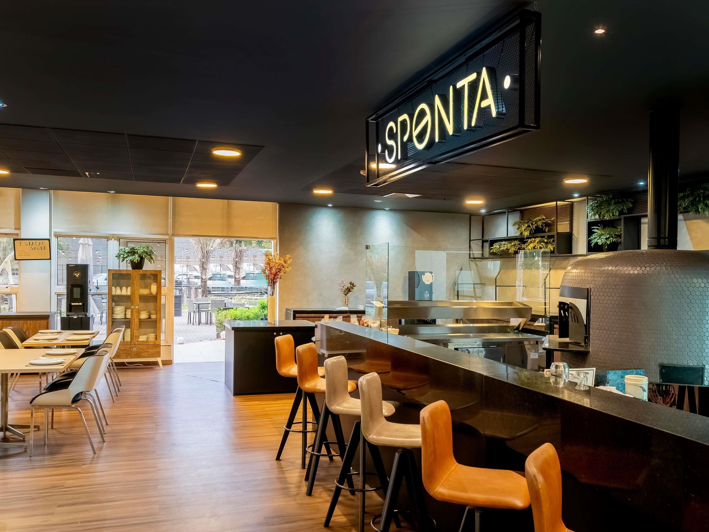 A modern bar area with a prominent 'SPONTA' sign, a counter with stools, and a view of the street outside.
