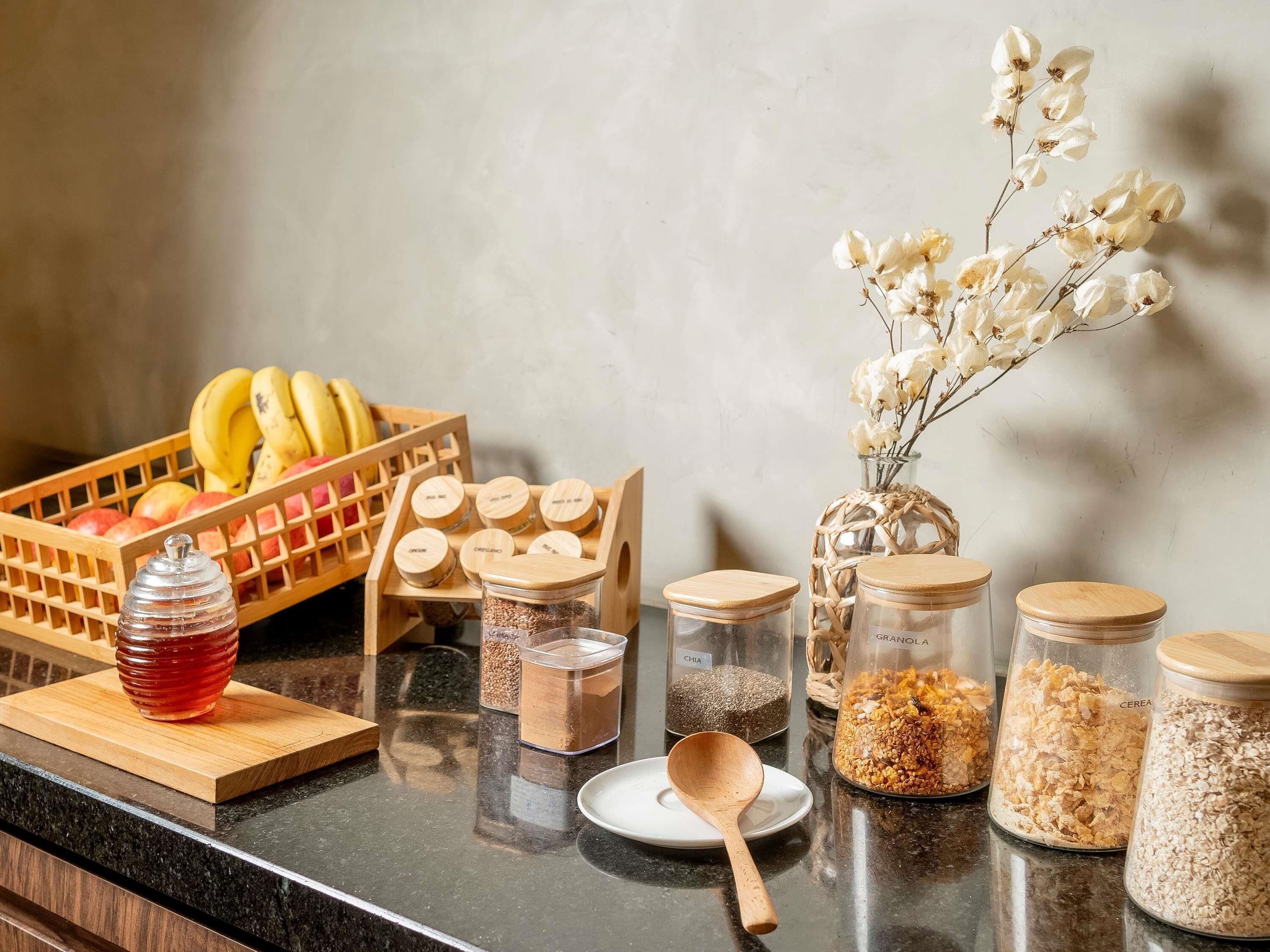 A kitchen counter with jars of grains, a honey jar, a fruit basket, and a decorative vase with dried flowers.