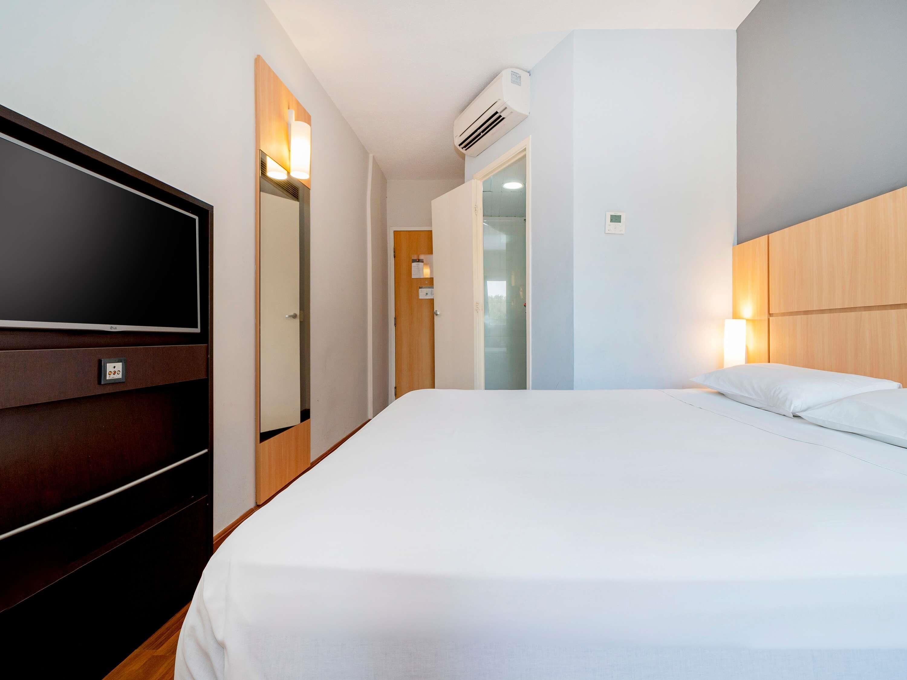Una habitación de hotel con una cama grande, un televisor de pantalla plana, un cabecero de madera y un aire acondicionado instalado en la pared.