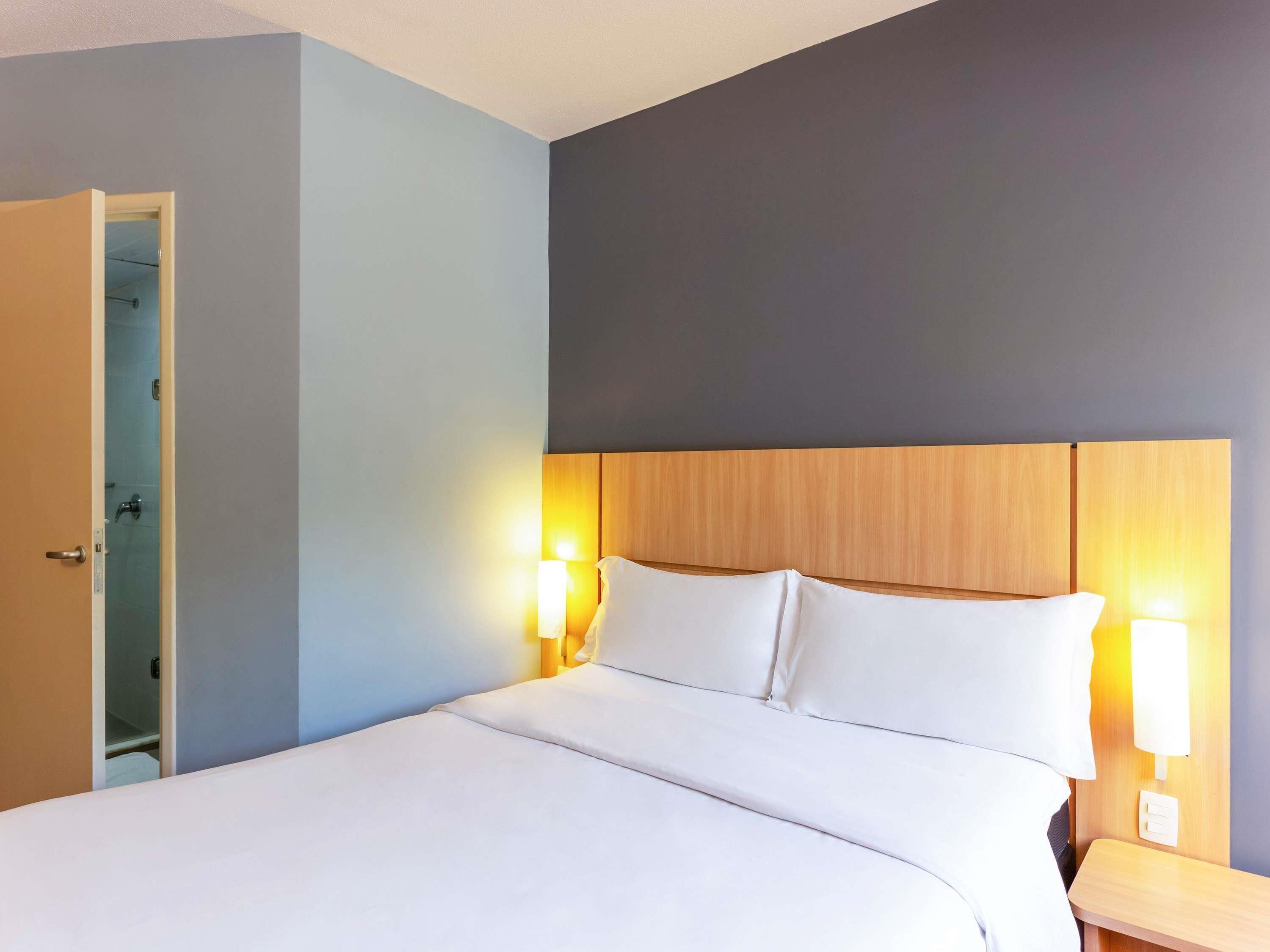 A hotel room with a bed, bedside lamps, a wooden headboard, and a door leading to a bathroom.