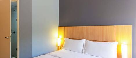 Habitación de hotel con cama, lámparas de noche, cabecera de madera y puerta que da a un baño.