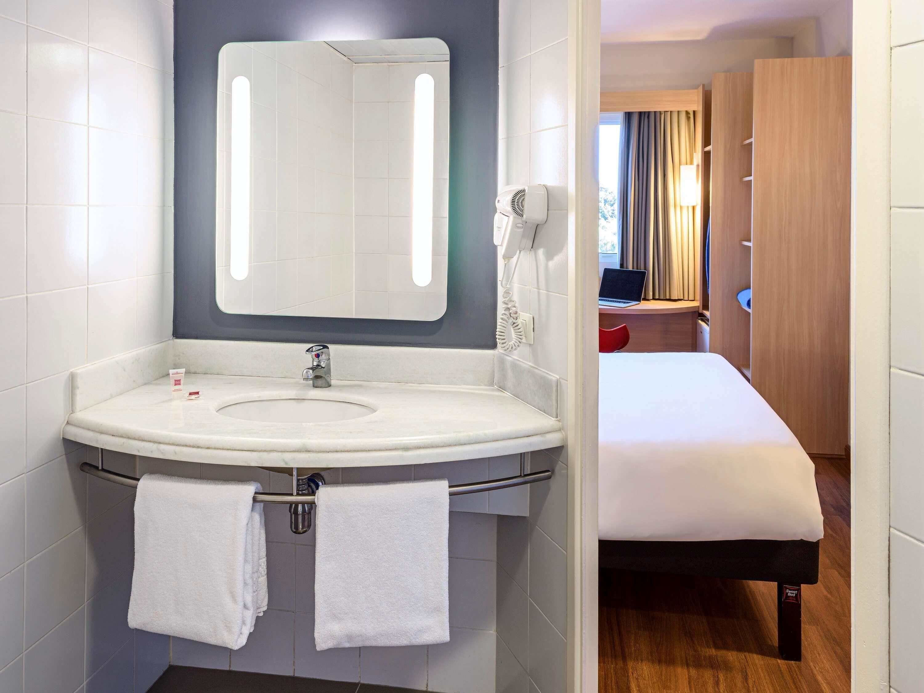 A hotel bathroom with a sink, mirror, towel rack, and a visible bed in the adjacent room.