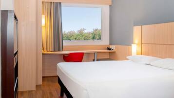 Habitación de hotel con cama, escritorio, silla, ventana con cortinas y vista a zonas verdes.