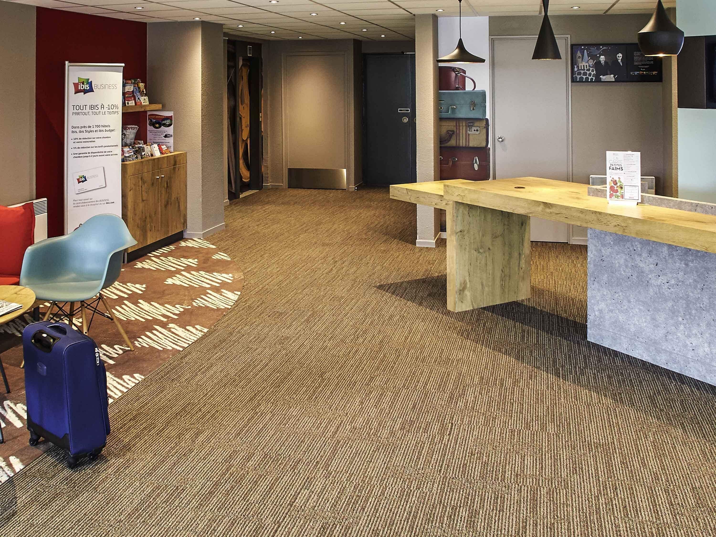A hotel lobby with a reception desk, a blue chair, a wooden table, and a luggage cart.