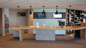 A reception area with a wooden reception desk, two televisions mounted on the wall, a bar area with shelves of bottles, and a storage unit with suitcases. - ibis Auray (Auray)