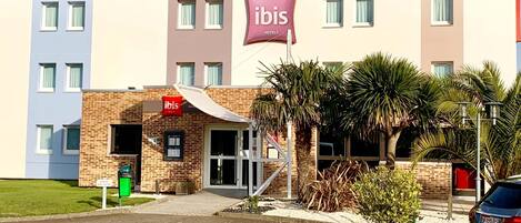 Um hotel com fachada moderna, um letreiro chamativo da 'ibis Hotels' e uma área de estacionamento com uma placa verde.