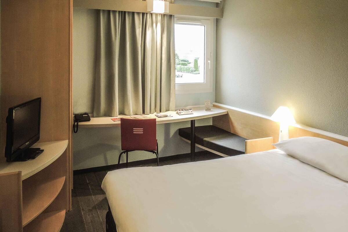 A hotel room with a bed, a desk with a chair, a TV, a window with curtains, and a lamp.