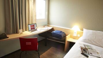 Une chambre d’hôtel équipée d’un bureau, d’un ordinateur portable, d’une chaise, d’une valise, d’une table de chevet avec une lampe, d’un lit et d’un magazine.