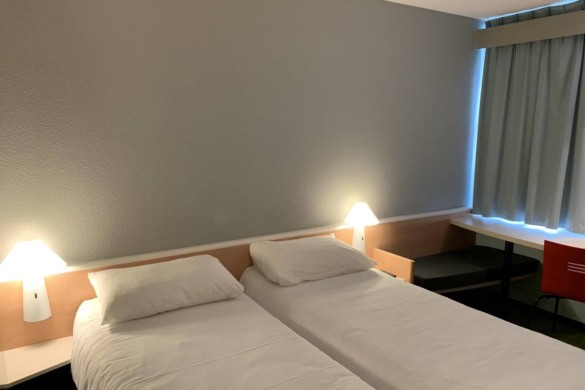 A hotel room with two single beds, a desk, a chair, and a window with curtains.