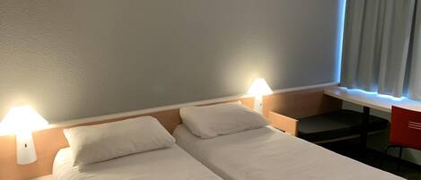 A hotel room with two single beds, a desk, a chair, and a window with curtains.