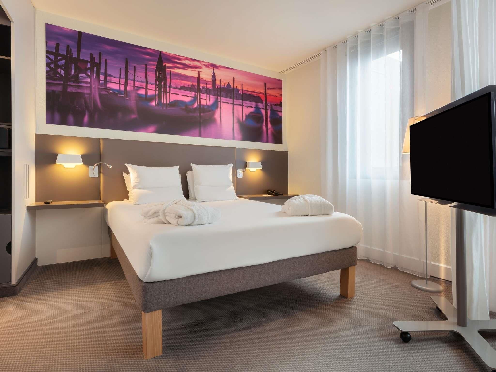 A hotel room with a bed, two bedside lamps, a flat-screen TV, and a large painting on the wall.