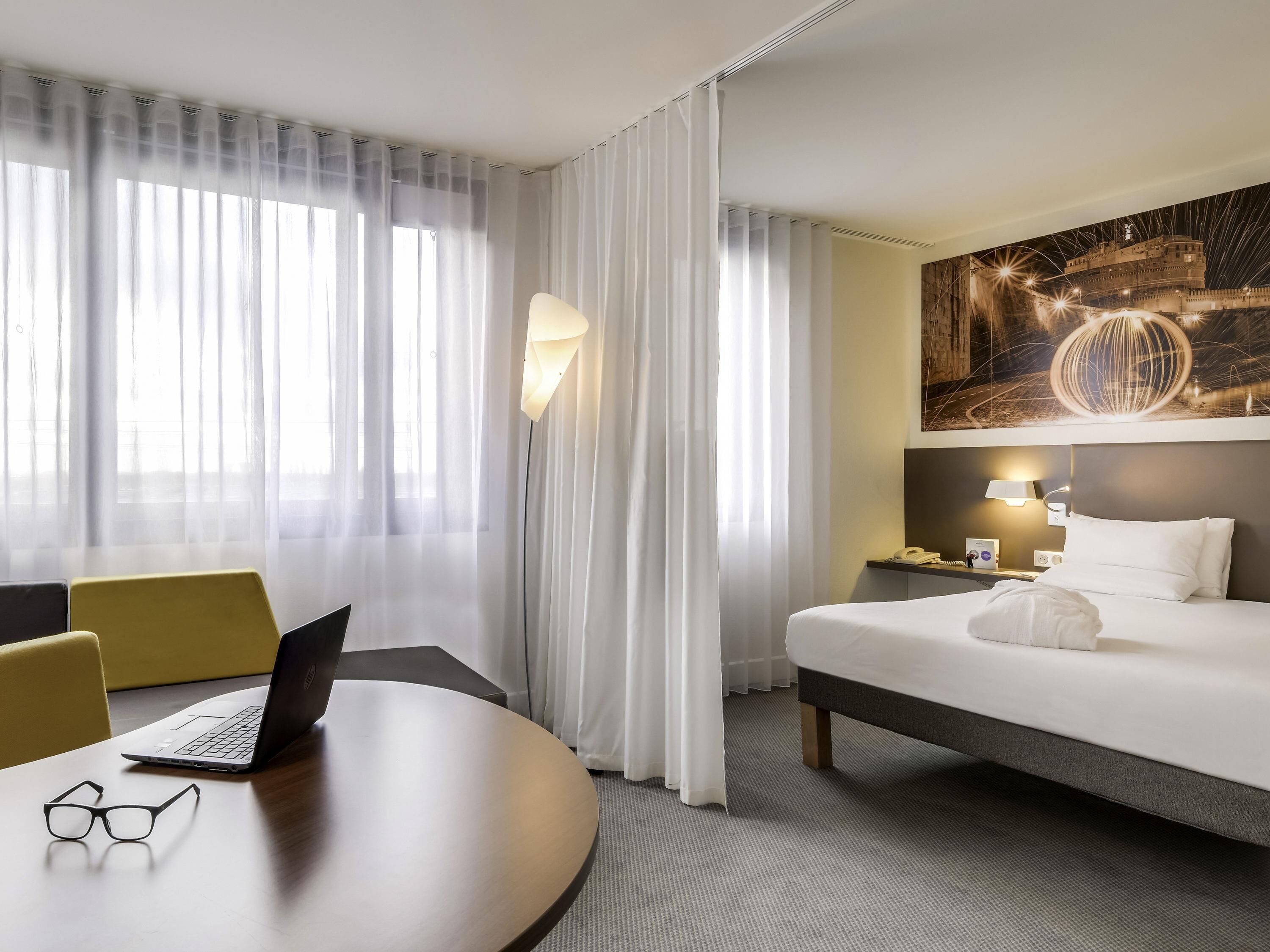 Une chambre d’hôtel avec un lit, un bureau avec un ordinateur portable et des lunettes, une table de chevet avec une lampe, et une grande fenêtre avec des rideaux légers.
