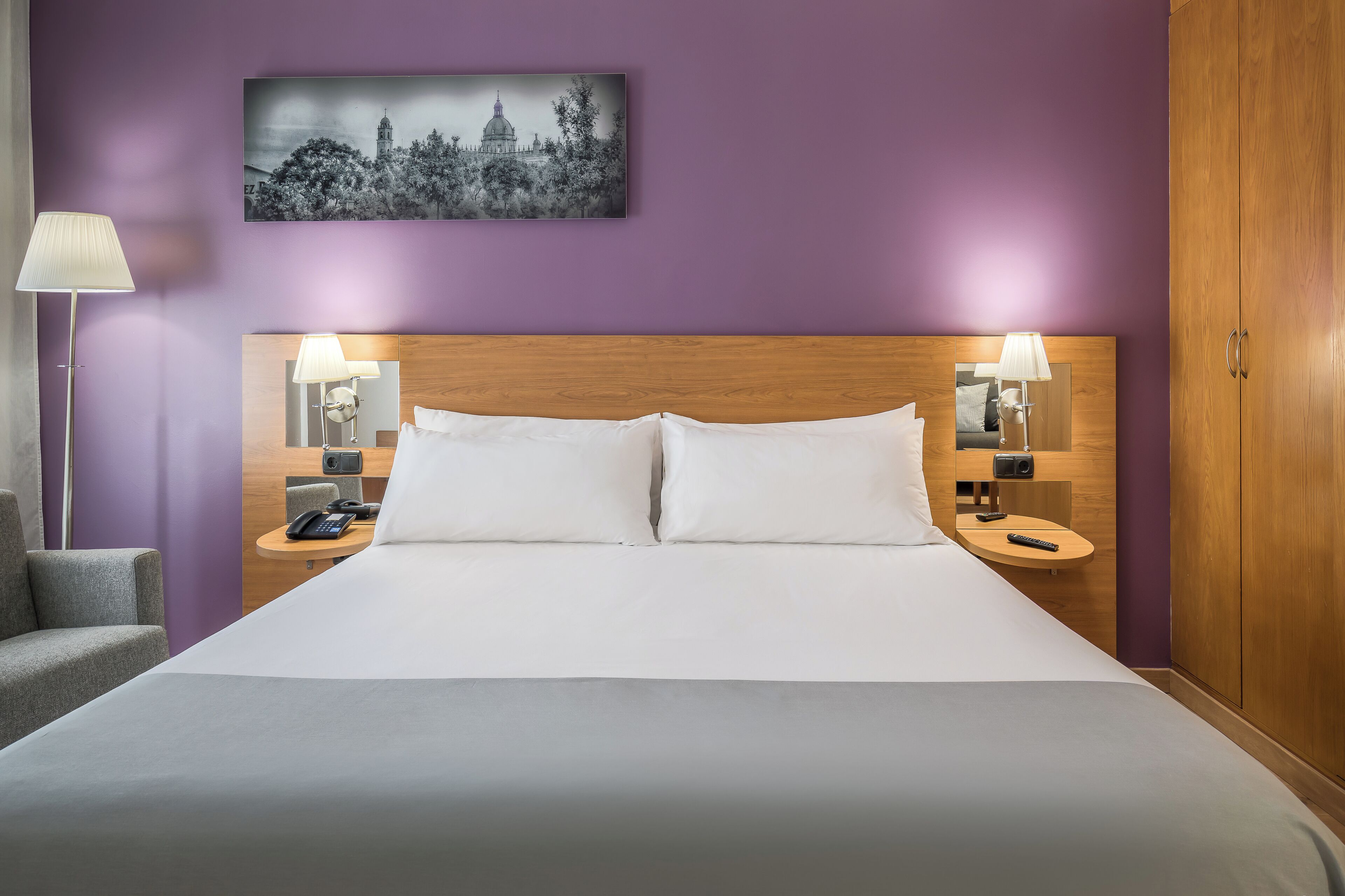 Een hotelkamer met een groot bed, twee nachtkastjes met lampen, een stoel en een aan de muur hangend schilderij.