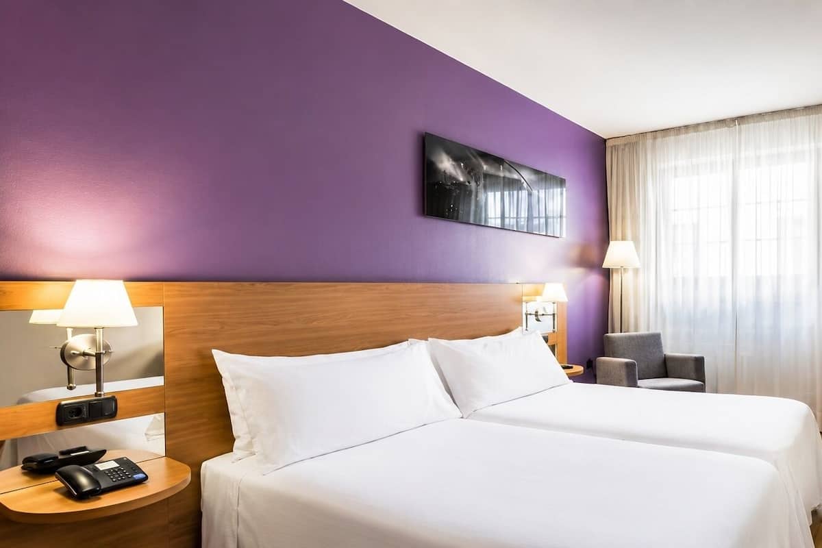 Une chambre d’hôtel comprenant un lit, deux tables de chevet, un bureau avec une chaise, une télévision et une fenêtre avec des rideaux.