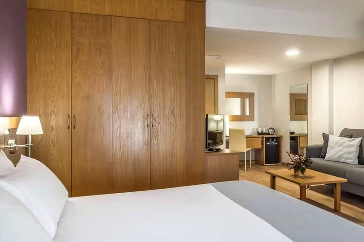 Une chambre d’hôtel comprenant un lit, une armoire en bois, un bureau avec une chaise, un canapé et un petit coin cuisine.