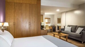 Habitación de hotel con cama, armario de madera, escritorio con silla, sofá y pequeña cocina.