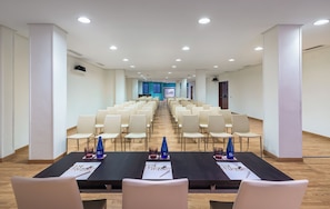 Un salón de conferencias con filas de sillas, una larga mesa con tarjetas de identificación y botellas de agua, y una pantalla para presentaciones al frente.