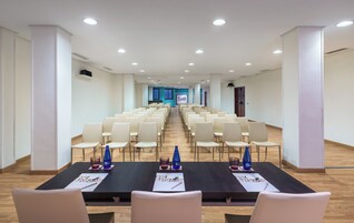 Un salón de conferencias con filas de sillas, una larga mesa con tarjetas de identificación y botellas de agua, y una pantalla para presentaciones al frente.