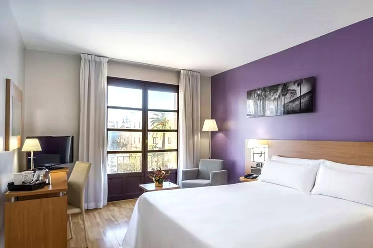 Une chambre d’hôtel avec un grand lit, un bureau avec une télévision, une chaise et une fenêtre avec des rideaux.