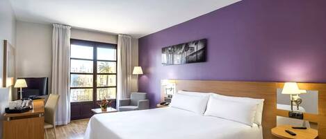 A hotel room with a large bed, a desk with a TV, a chair, and a window with curtains.