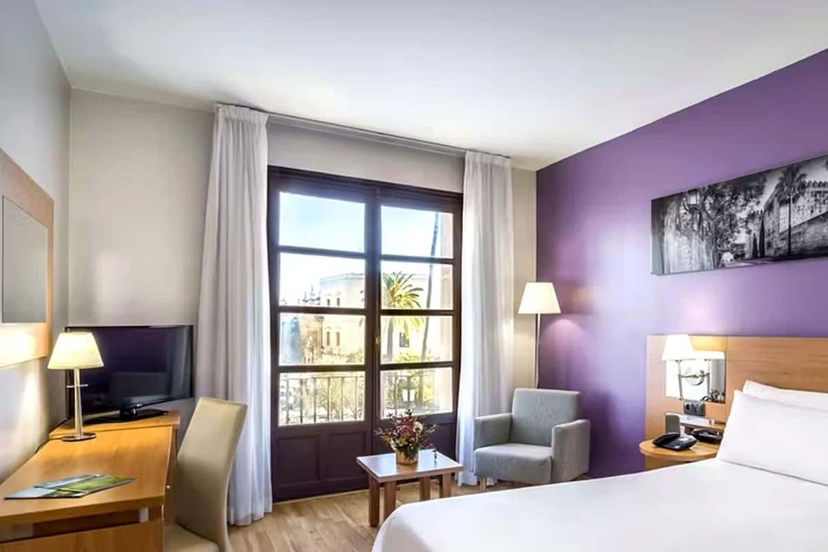 Une chambre d’hôtel avec un lit, un bureau, une chaise, une télévision et une fenêtre avec des rideaux.