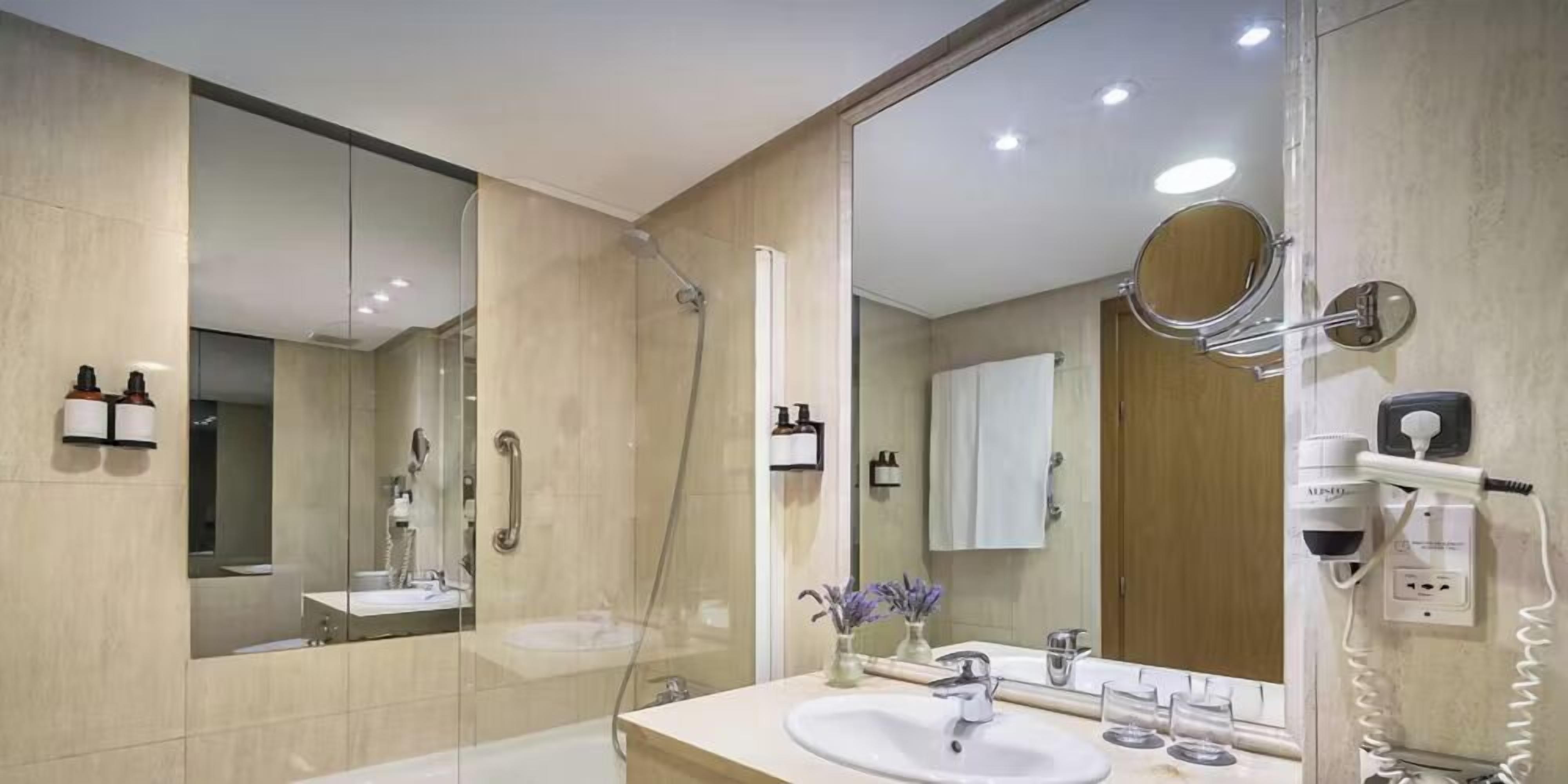 A modern bathroom with a large mirror, two sinks, a shower, and a hairdryer on the wall.
