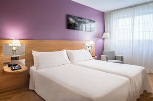 Habitación de hotel con dos camas, cabecero de madera, mesita de noche con teléfono, una silla y ventana con cortinas translúcidas.