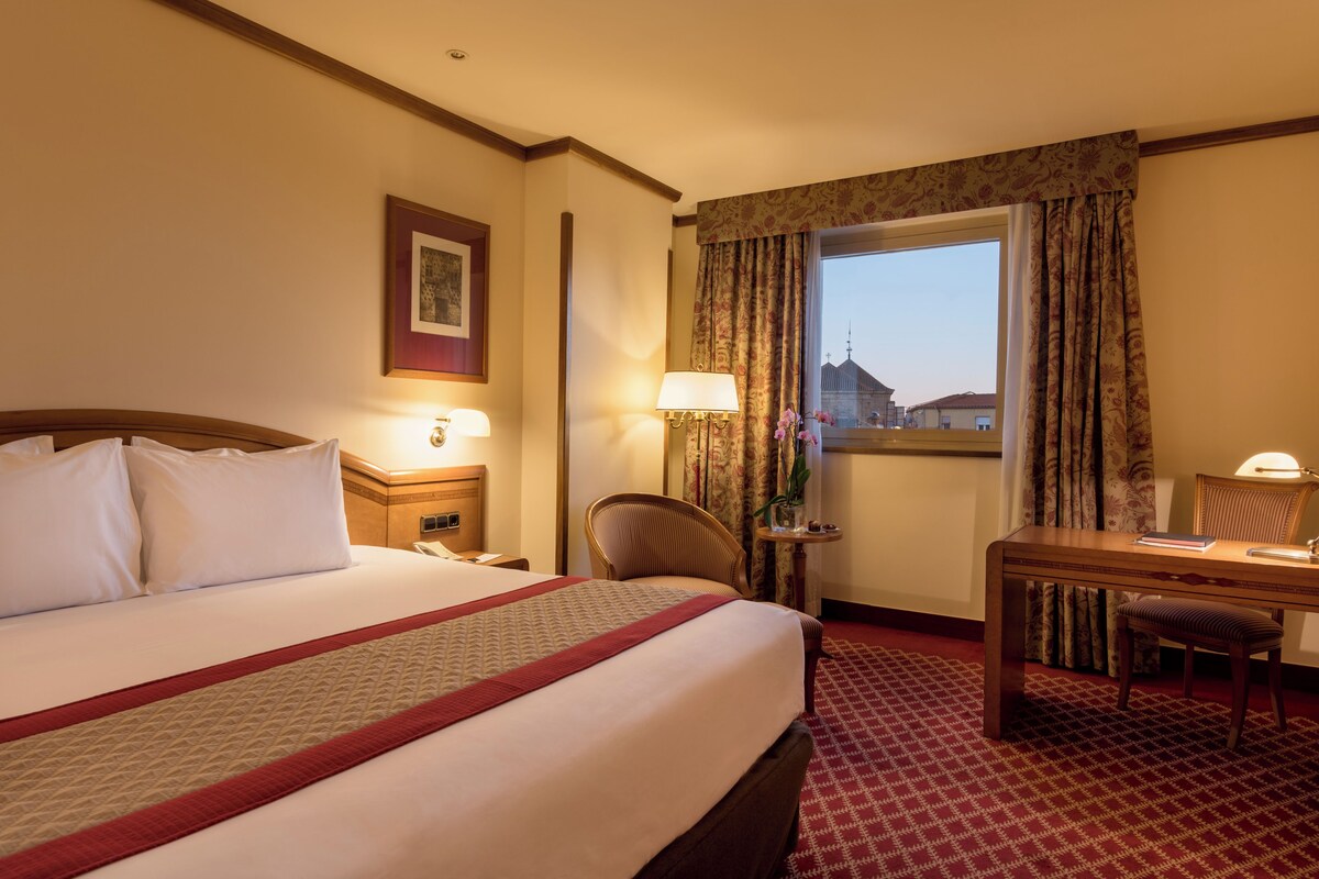 Een hotelkamer met een groot bed, een bureau, een stoel, een lamp en uitzicht op een gebouw.