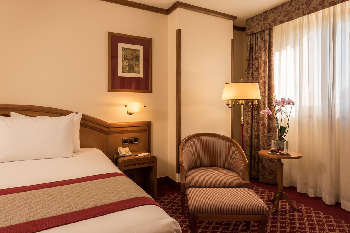 Een hotelkamer met een groot bed, een nachtkastje met een lamp, een stoel en een tafeltje met een bloevas.