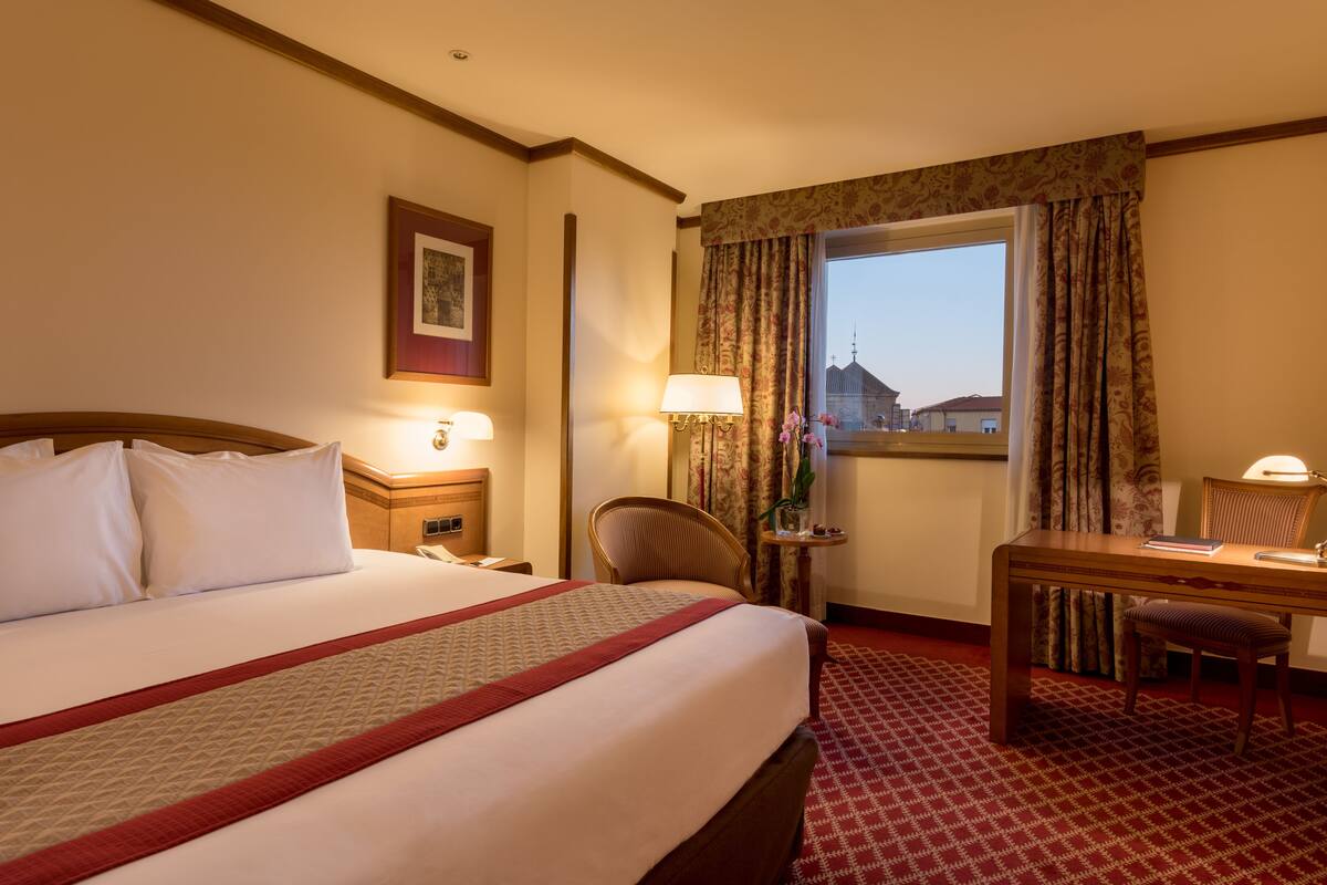 Een hotelkamer met een groot bed, een bureau, een stoel, een lamp en een raam met gordijnen.