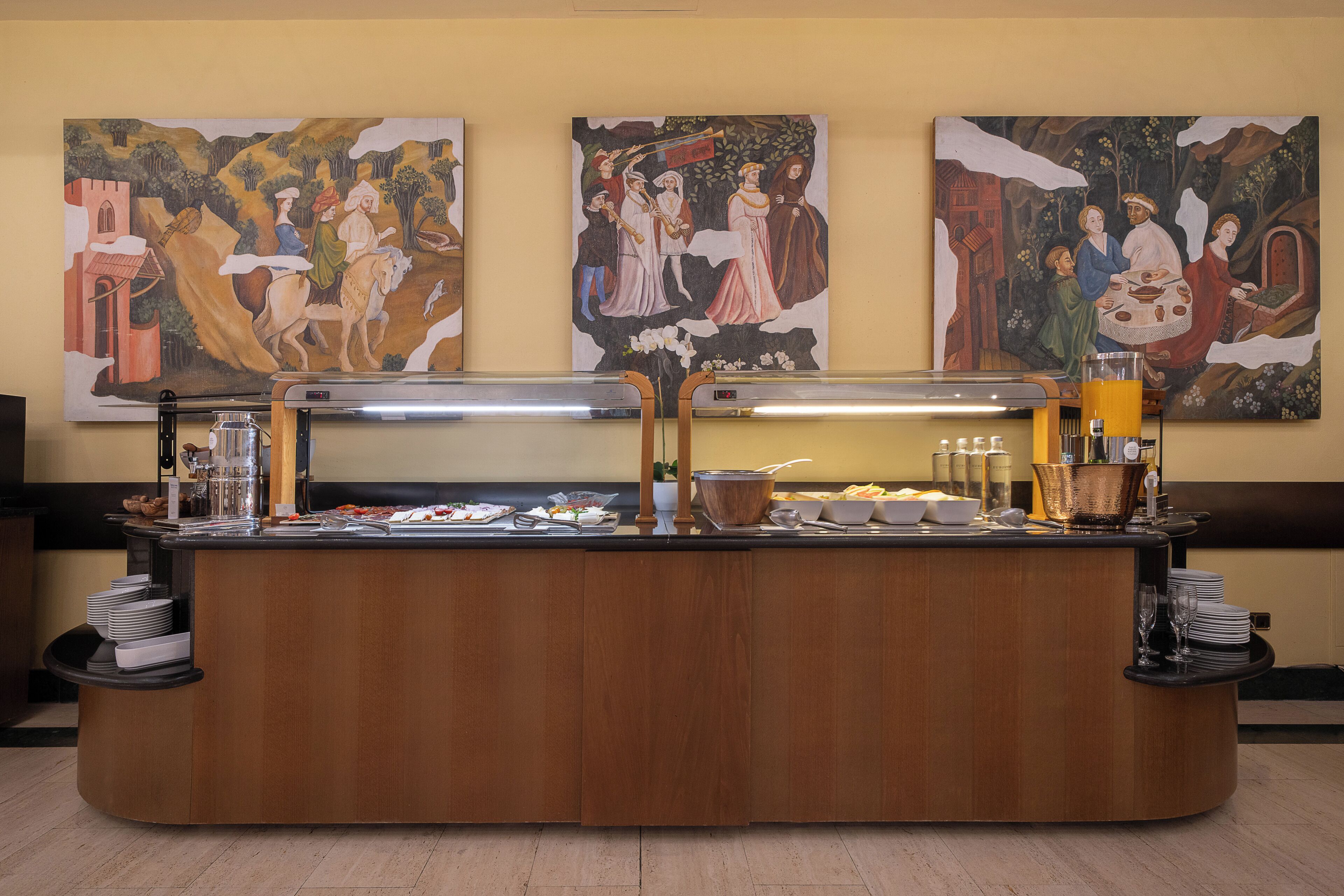 A buffet counter with a variety of food items, a coffee station, and three large paintings on the wall.