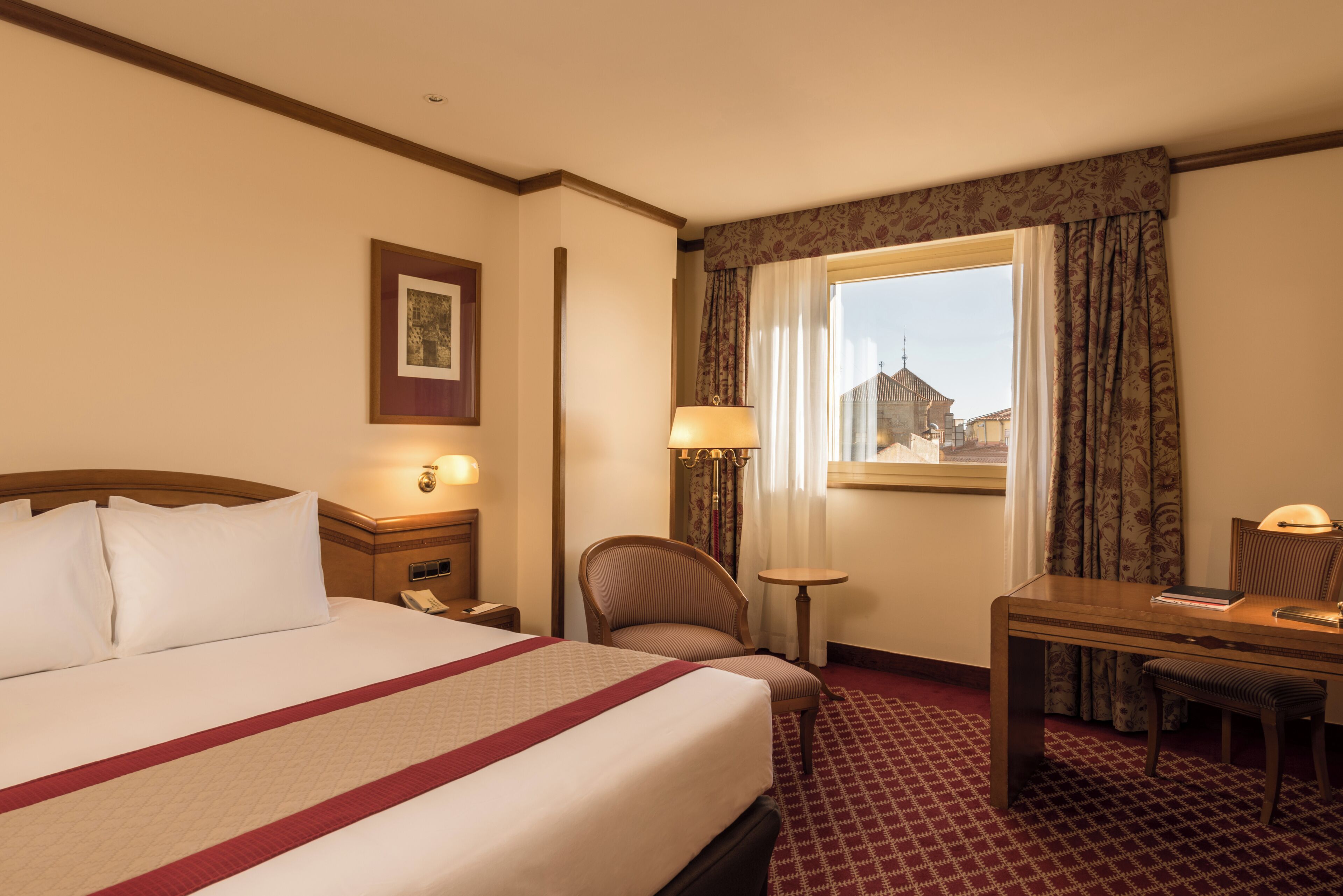 Une chambre d’hôtel avec un lit, un bureau, une chaise, une fenêtre avec des rideaux et une lampe.