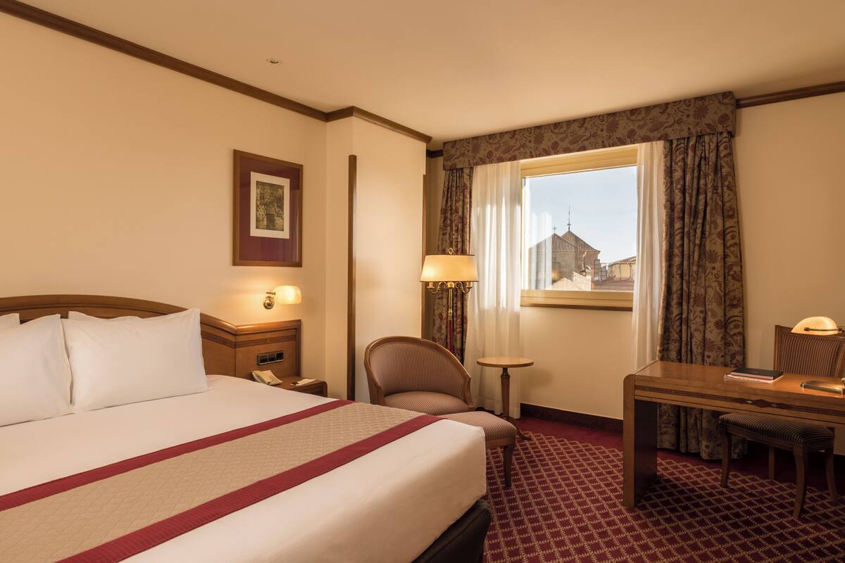 Een hotelkamer met een bed, een bureau, een stoel, een raam met gordijnen en een lamp.