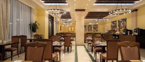 A hotel dining area with multiple tables and chairs, a central column, and a kitchen area in the background.