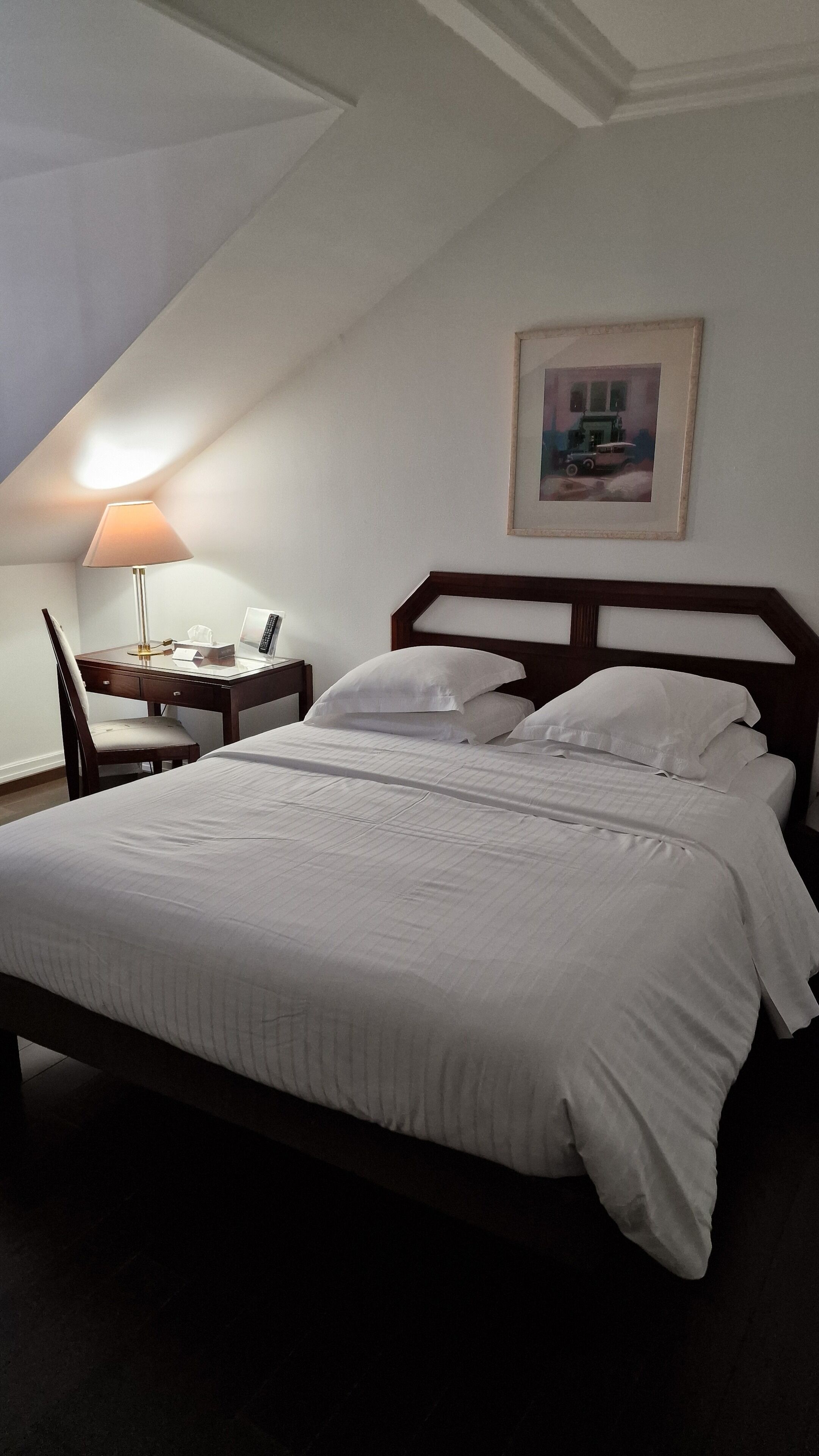 A neatly made bed with white linens in a room with a sloped ceiling. There is a desk with a lamp and a telephone, and a framed picture on the wall.