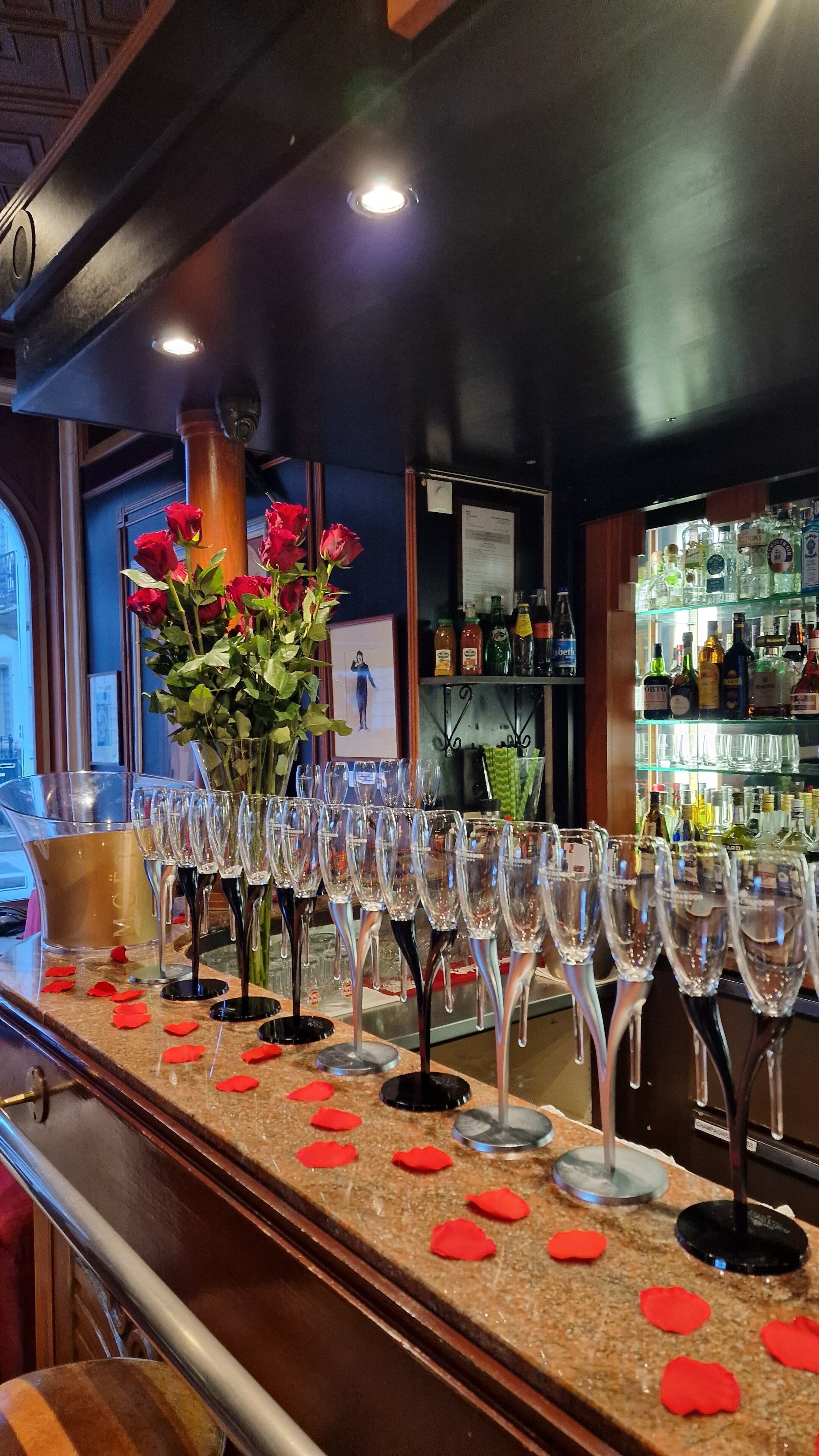 A bar area with a long counter, multiple wine glasses, and a vase of red roses.